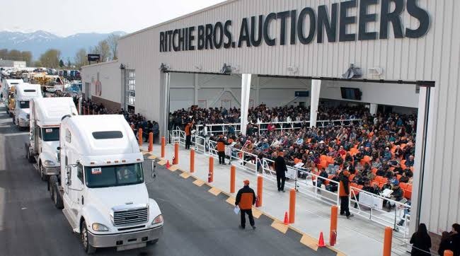 Large live auction event with crowd of bidders and equipment lined up outside the facility
