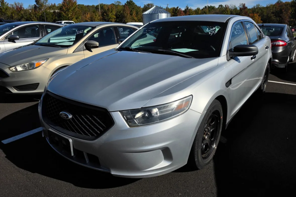 A silver Ford Taurus sedan with black steel wheels in the Delaware Surplus Services yard, with a Ford Focus beside it.