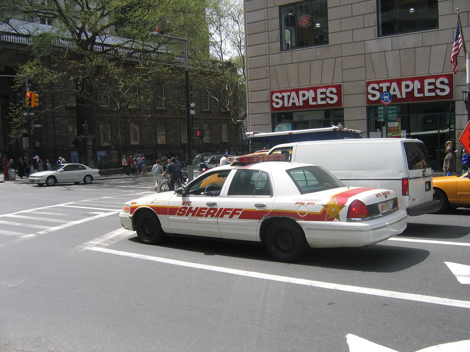 A white New York City Sheriff Crown Victoria with red and yellow striping, turning at a Manhattan intersection.