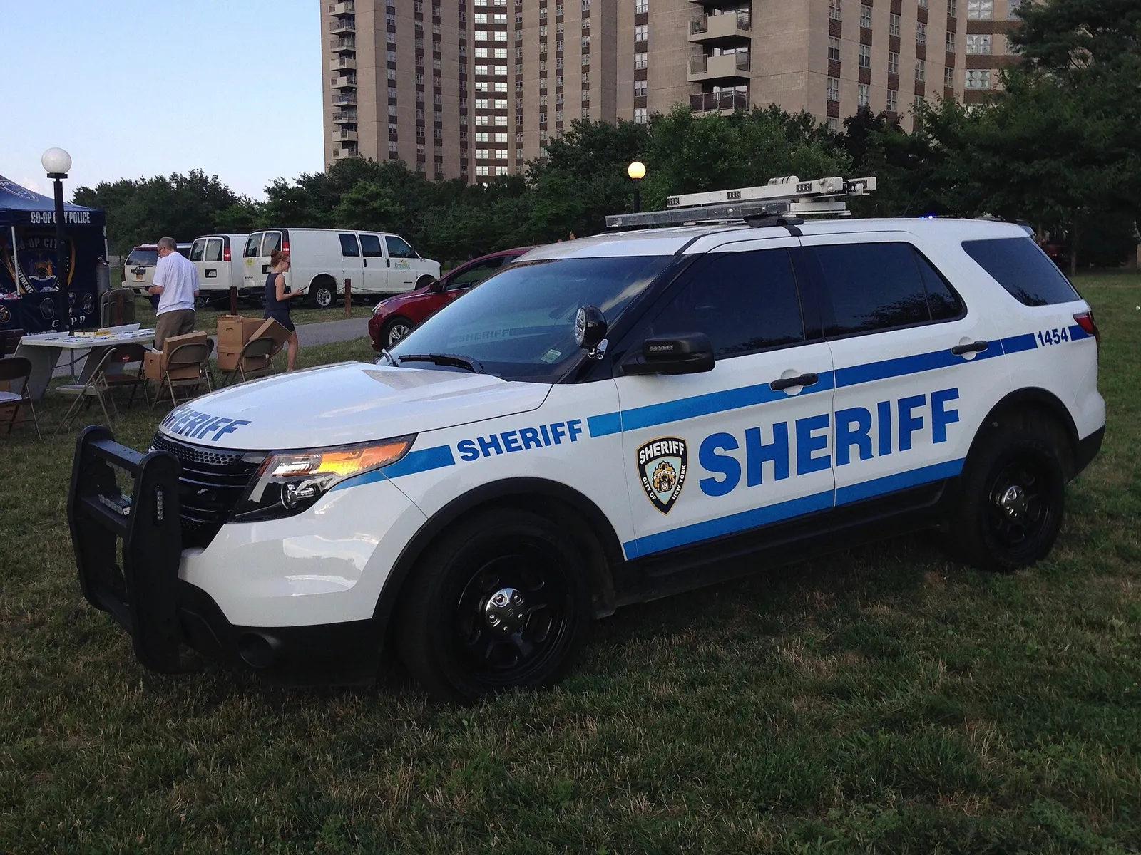 A white New York City Sheriff Ford Police Interceptor Utility parked on grass at a community event, with a Bronx apartment tower behind it.